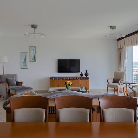 Hotel Grand Hyatt Dubai Esterno foto The photo shows a modern living room with a spacious and minimalistic design. There is a flat-screen television mounted on the wall, facing a seating arrangement that includes two armchairs and a sofa. A wooden table is positioned in the foreground, 