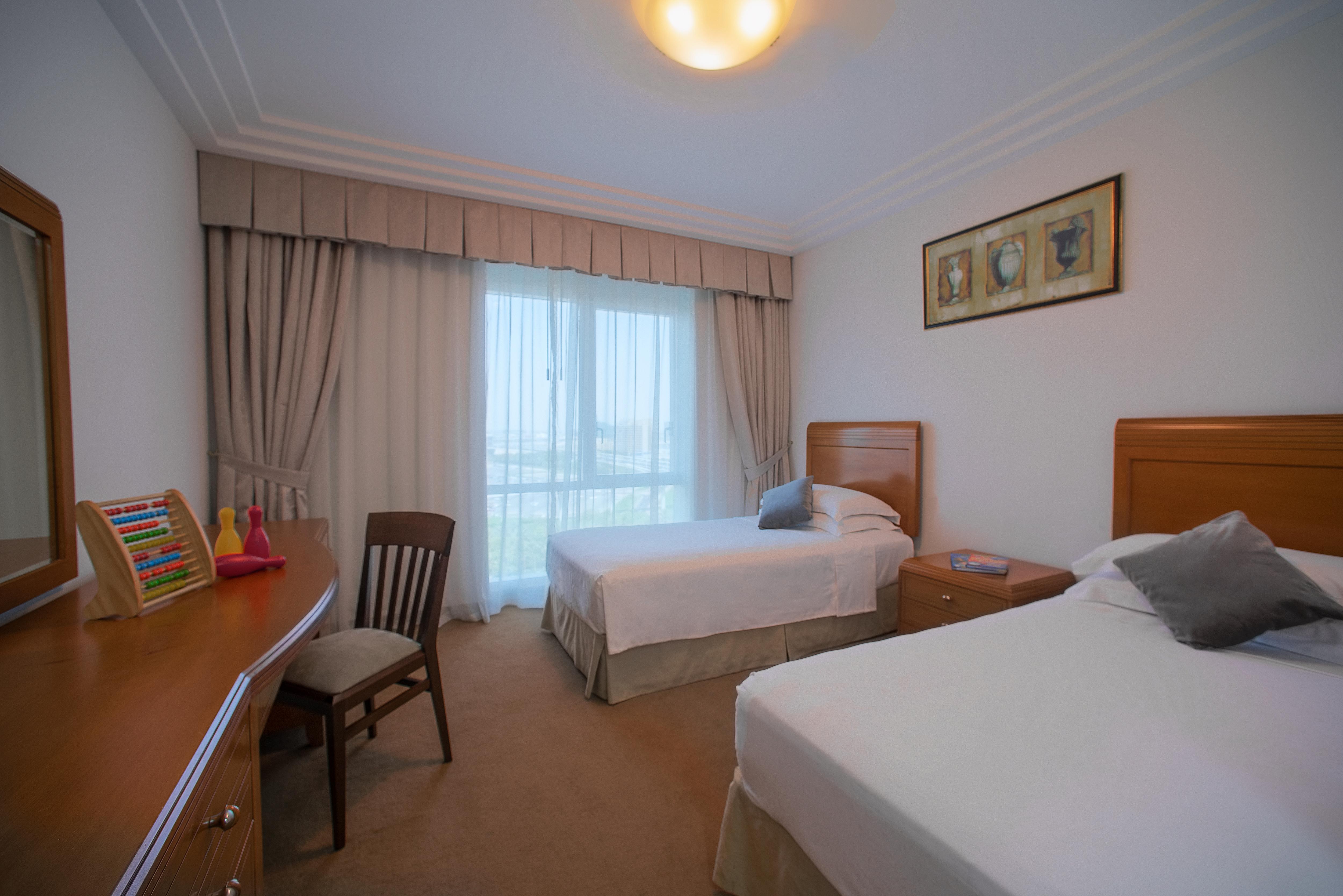 Hotel Grand Hyatt Dubai Esterno foto The photo shows a spacious hotel room featuring two single beds with white bedding, positioned against a wall. There is a wooden dresser next to the bed, along with a chair. A large window, covered with light-colored curtains, allows natural light to