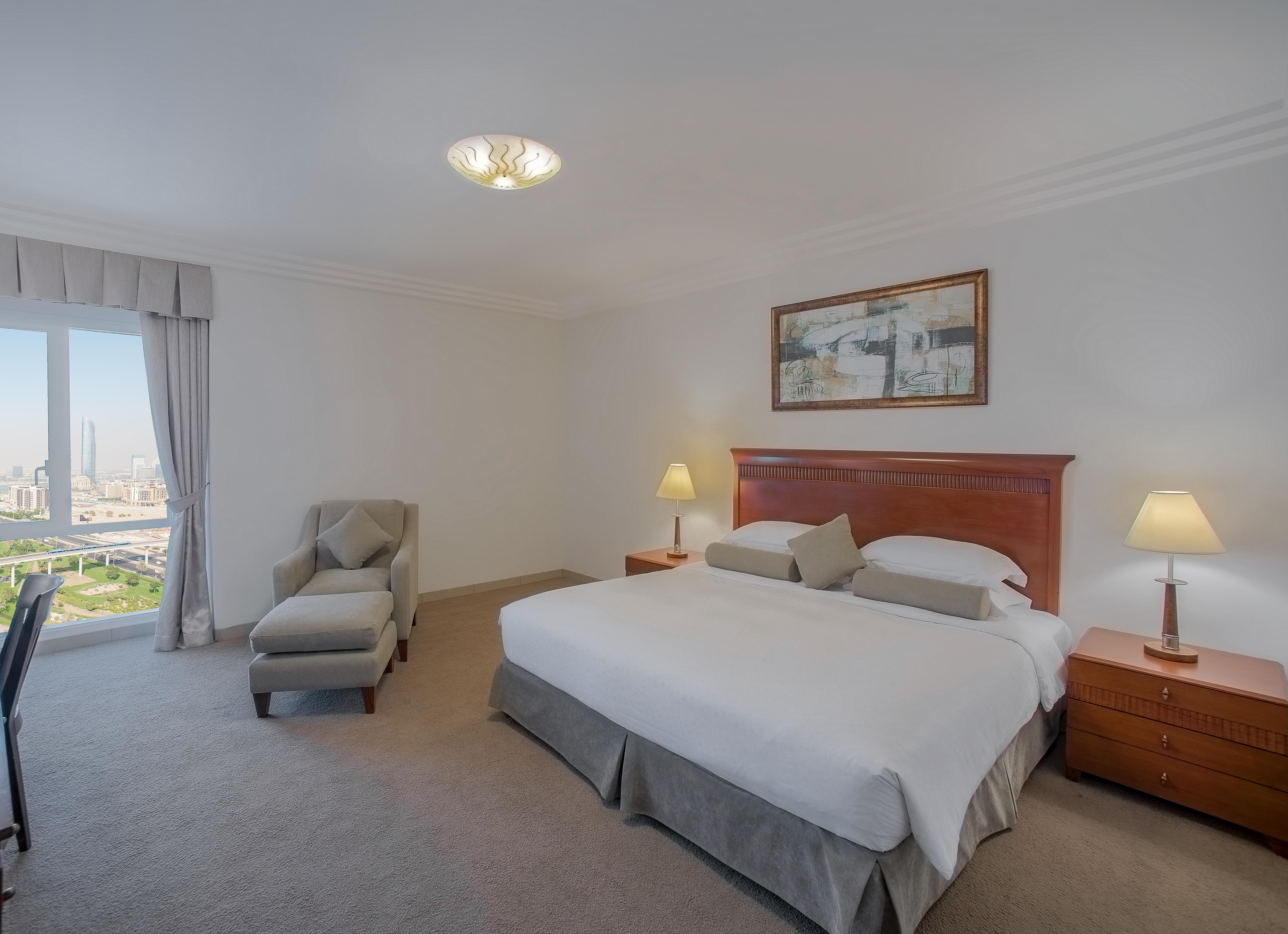 Hotel Grand Hyatt Dubai Esterno foto The photo shows a spacious hotel room with a contemporary design. There is a large bed with white linens and two decorative pillows. Next to the bed are two bedside tables with lamps. On one wall, there is a framed artwork. A comfortable armchair and