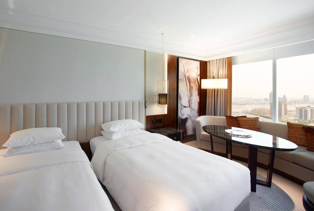 Hotel Grand Hyatt Dubai Esterno foto The photo shows a modern hotel room featuring two separate beds with white bedding. The walls are painted in neutral tones, and there is a large window that allows natural light to enter, offering a view of the city. A stylish round table and chairs 