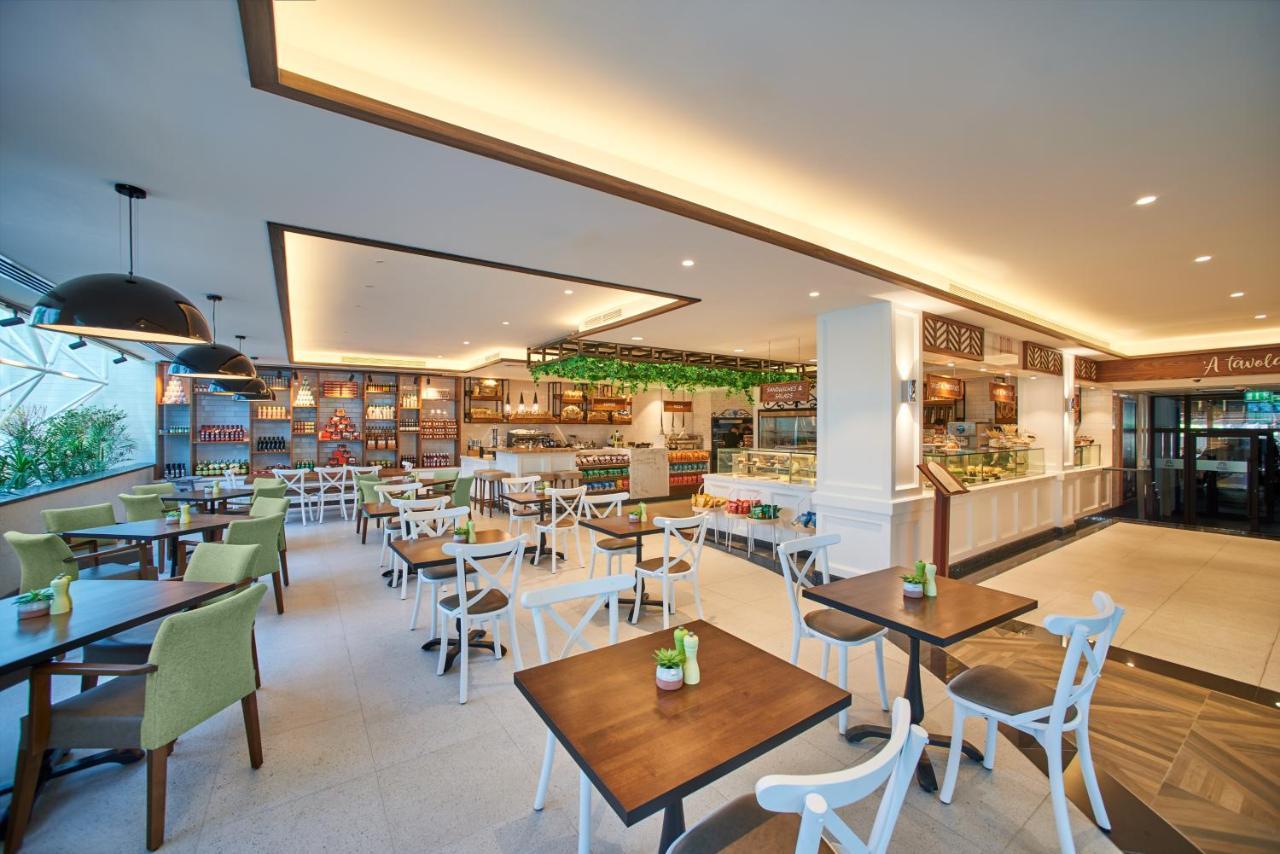 Hotel Grand Hyatt Dubai Esterno foto The photo shows a modern café or restaurant interior. It features a spacious layout with wooden tables and chairs, some of which have white frames and green cushions. The walls are adorned with shelves showcasing various items, and there is a display