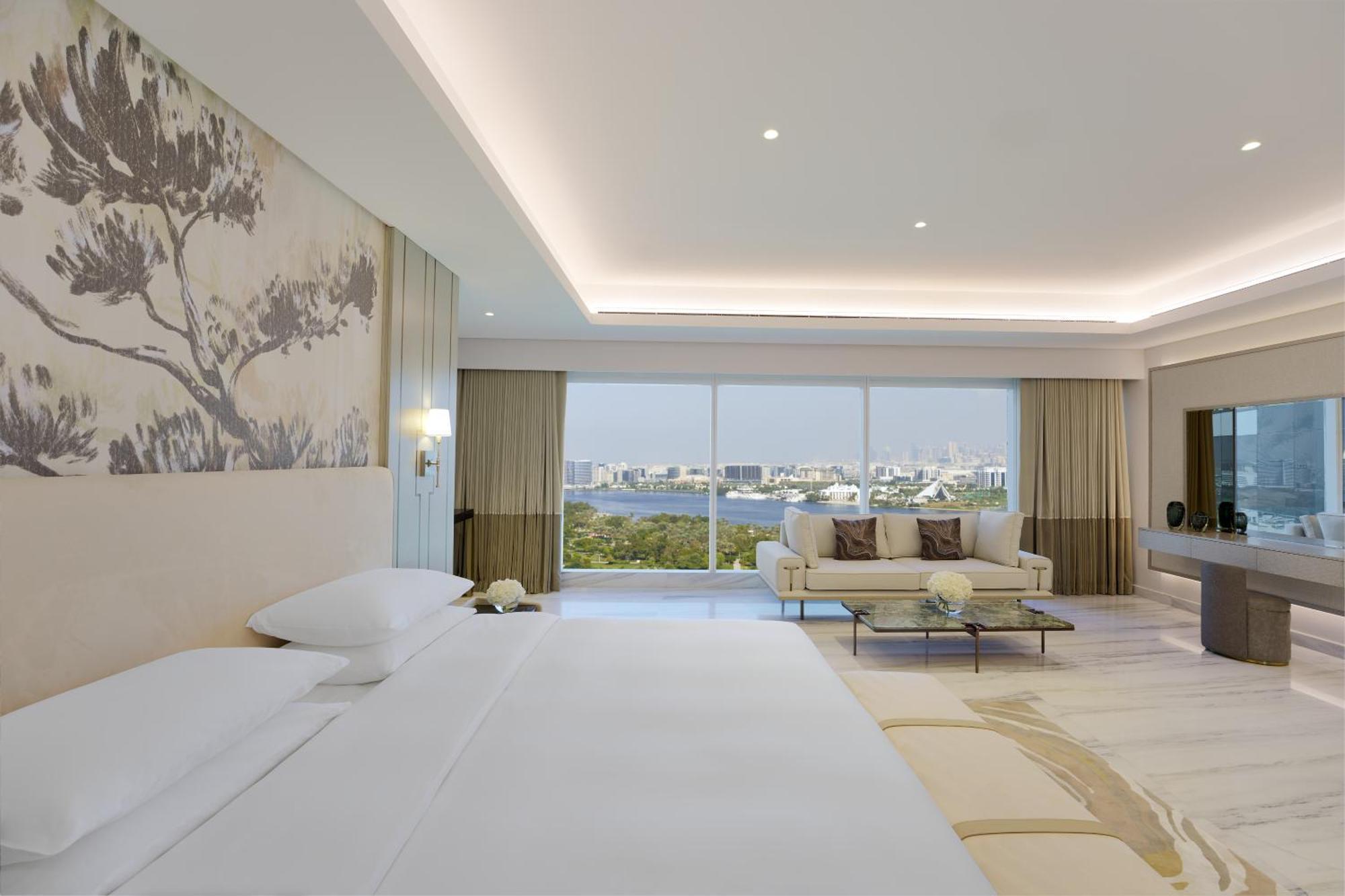 Hotel Grand Hyatt Dubai Esterno foto The photo shows a modern, elegantly designed hotel room. It features a large bed with white linens, positioned against a beautifully decorated wall with a subtle nature-themed mural. There is a cozy seating area with a light-colored sofa and decorati