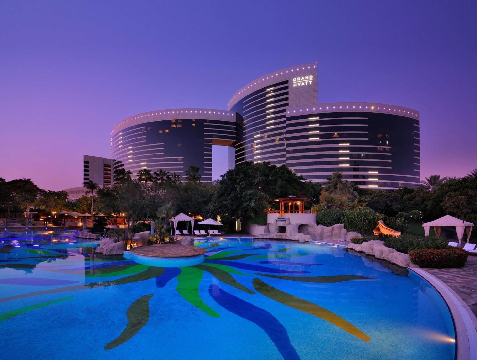 Hotel Grand Hyatt Dubai Esterno foto The photo shows a luxurious hotel complex set against a twilight sky. The architecture features modern, curved buildings with large windows, reflecting the evening light. In the foreground, there is an expansive pool featuring colorful patterns on th