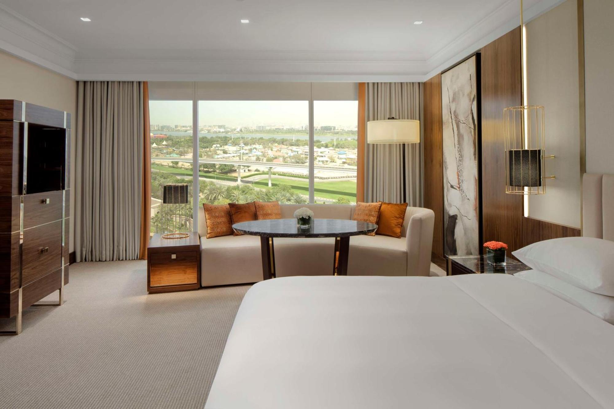 Hotel Grand Hyatt Dubai Esterno foto The photo shows a modern hotel room. In the foreground, there is a comfortable bed with a white bedspread. To the side, you can see a cozy sitting area with a round table and a couch, adorned with orange cushions. Behind the seating area, large windo