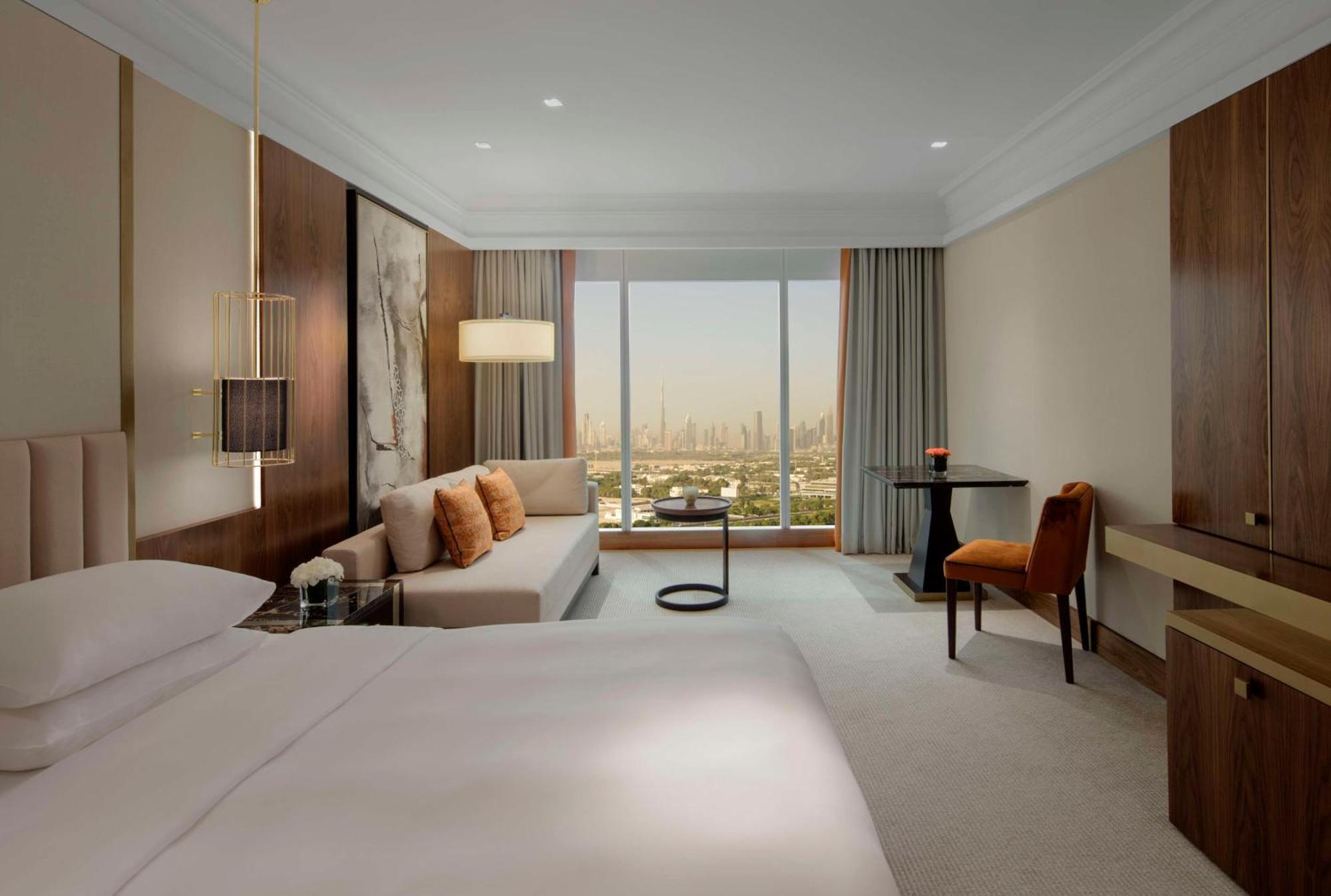 Hotel Grand Hyatt Dubai Esterno foto The photo shows a modern hotel room with a spacious design. It features a large bed with white linens, a comfortable couch adorned with pillows, and a small round table. A stylish chair sits next to a desk or dresser. The room has large windows offer